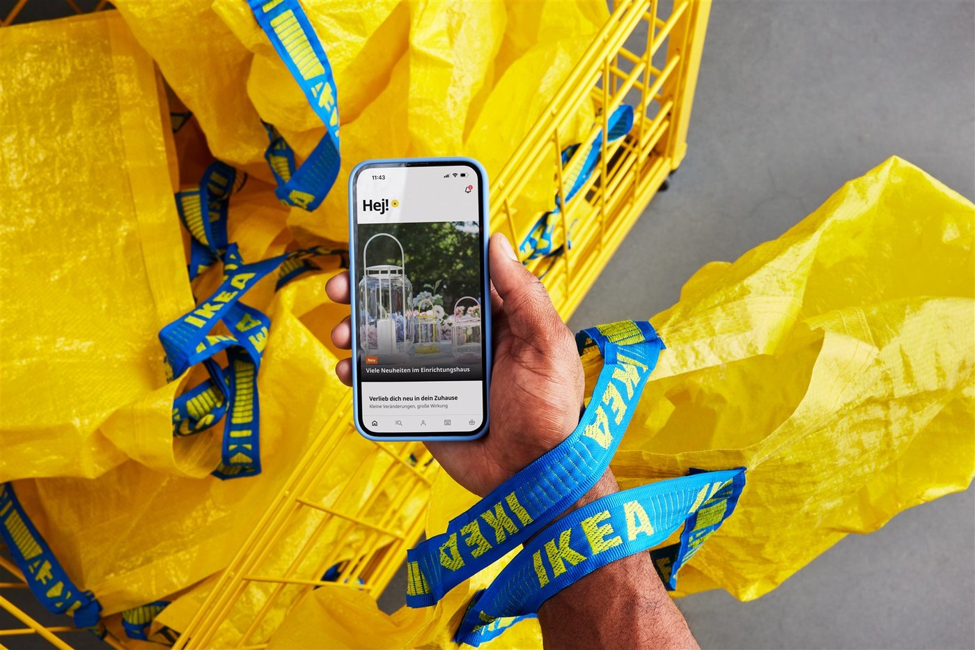 Eine Person hält Handy mit einer geöffneten IKEA App im IKEA Einrichtungshaus vor gelben FRAKTA Taschen