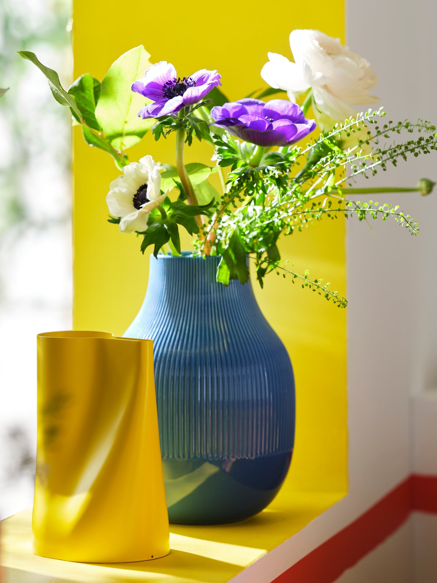 CHILIFRUKT yellow watering can standing next to a blue GRADVIS vase with flowers in it on a window shelf.