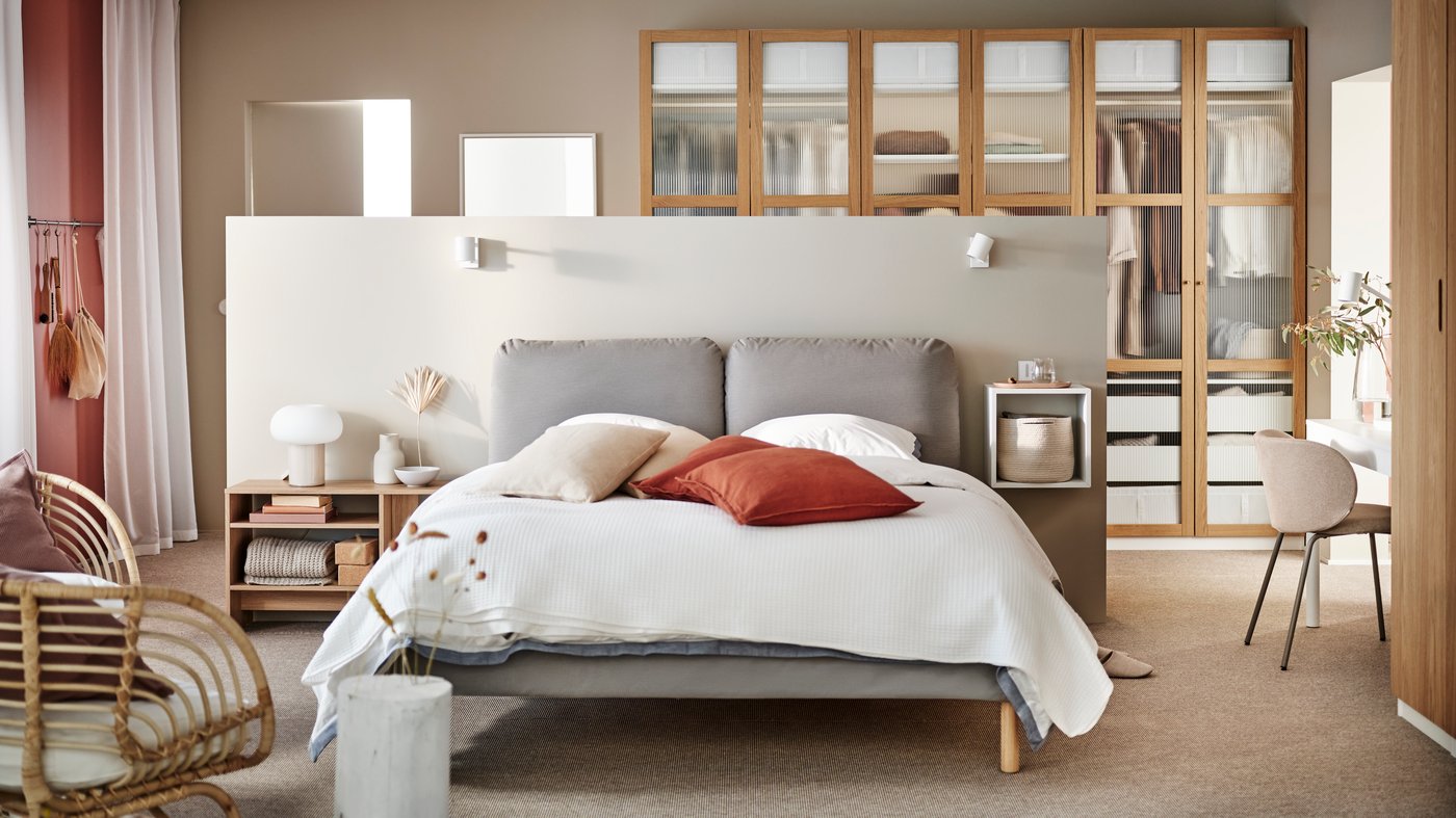 SAGESUND gestoffeerd bed in Diseröd bruin/Luröy, opgemaakt in het wit en met rode kussens erop, in een grote, luchtige slaapkamer.