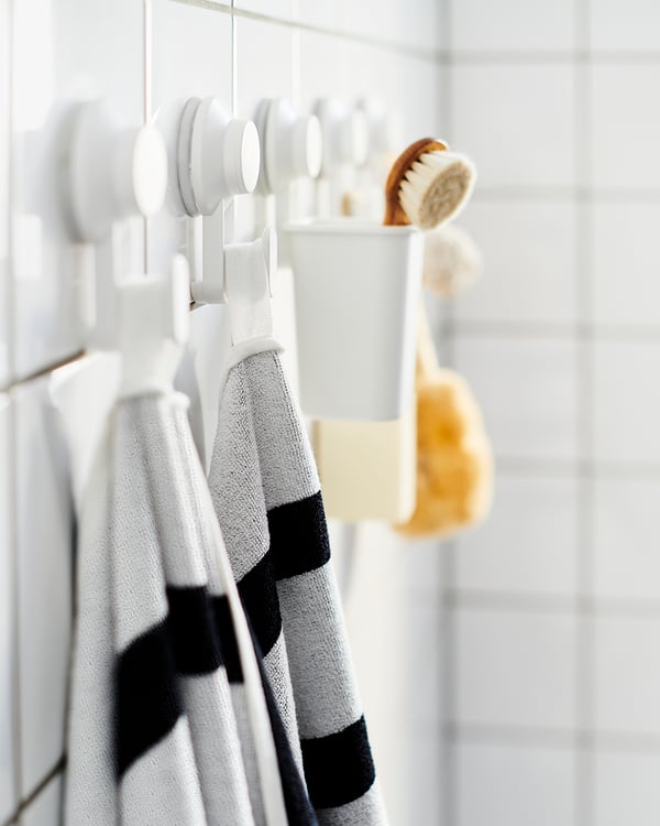 Cinco ganchos con ventosas TISKEN blancos colgados en fila en la pared de un baño con toallas de rayas, esponjas y cepillos.