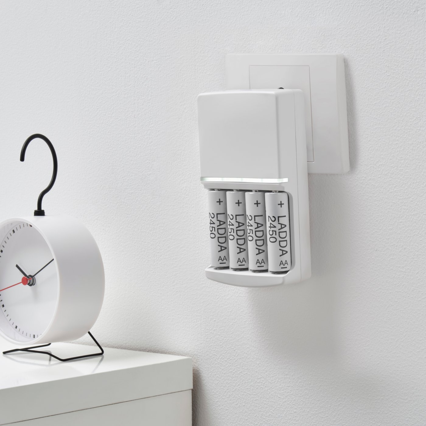 A white wall with LADDA recharcheable batteries charging in a KVARTS battery charger next to a clock on a white side table.