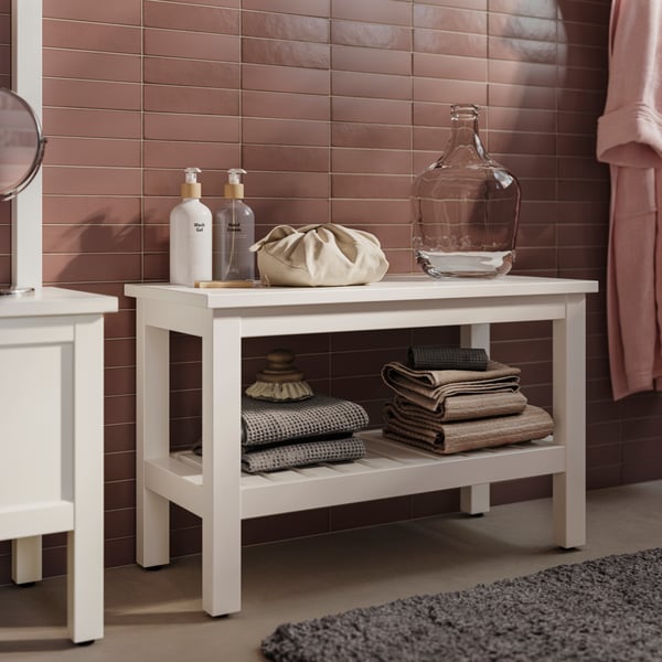 A bathroom with a HEMNES bench in white holding VINARN bath towels in light grey beige and an ALMTJÄRN bath mat in beige.