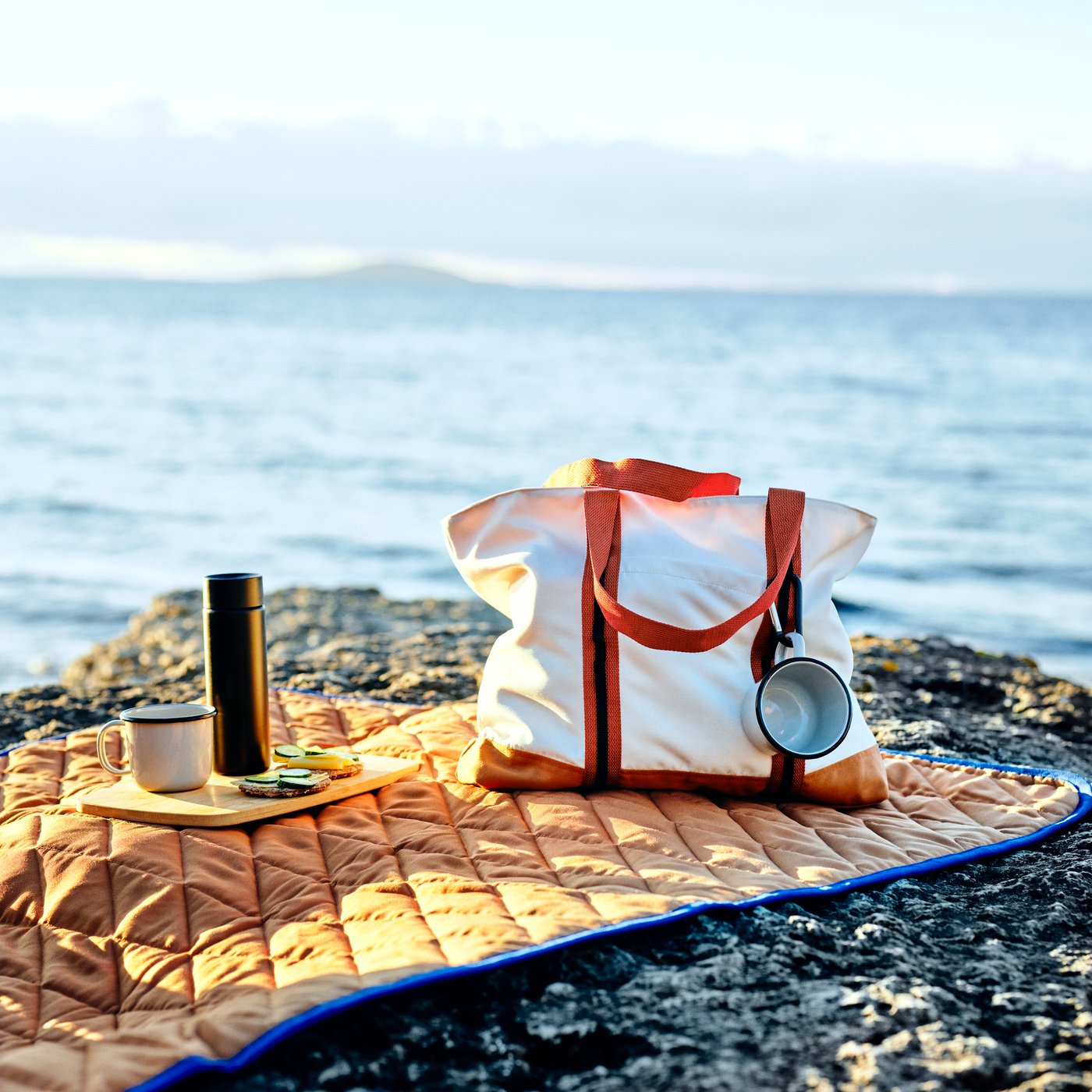 Eine SOLUPPGÅNG-Tasche, zwei Becher und ein Schneidebrett liegen auf einer SOLUPPGÅNG-Picknickdecke auf einer felsigen Klippe am Meer.