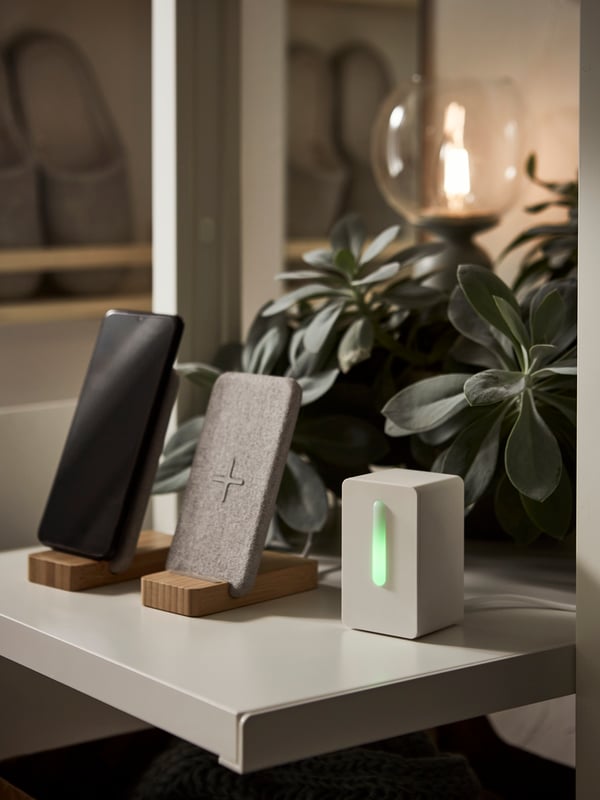 A VINDRIKTNING air quality sensor and two NORDMÄRKE wireless charging stands on a shelf with a lush plant behind them.