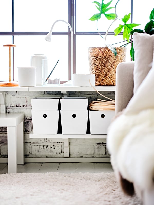 Two levels of white BERGSHULT/RAMSHULT wall shelves with white KUGGIS boxes with lids and a white NÄVLINGE LED work lamp.