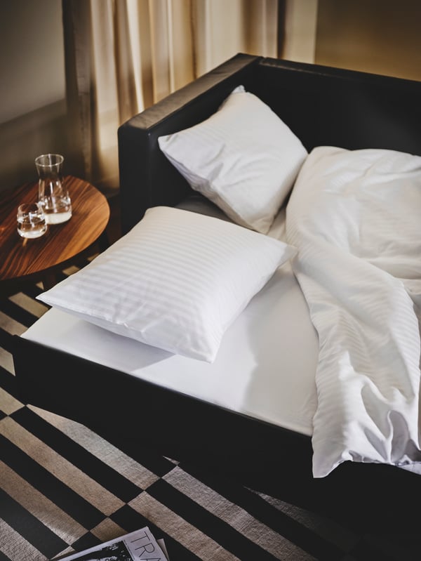 A black leather LANDSKRONA sofa is pulled out into a bed. A soft white duvet and two soft white pillows lie on top.