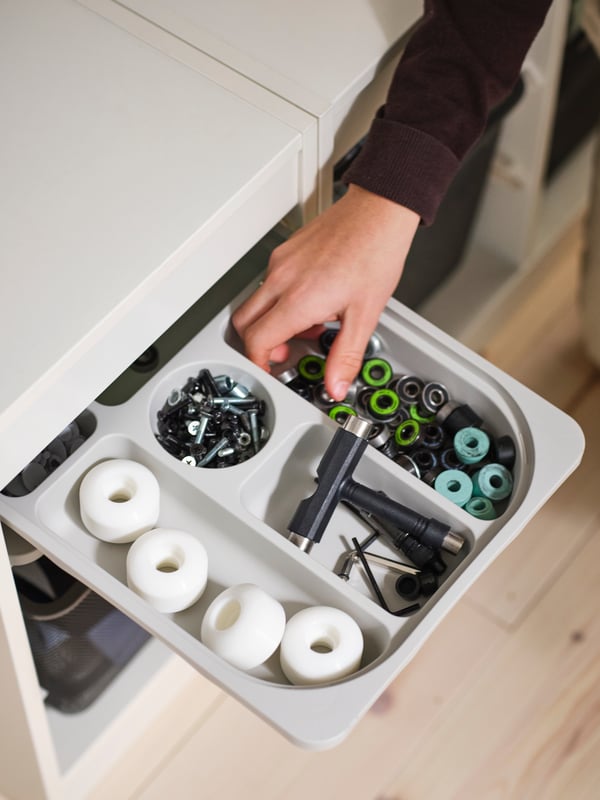 A young person’s hand reaching into a compartment in a light grey TROFAST storage tray holding skateboarding equipment.