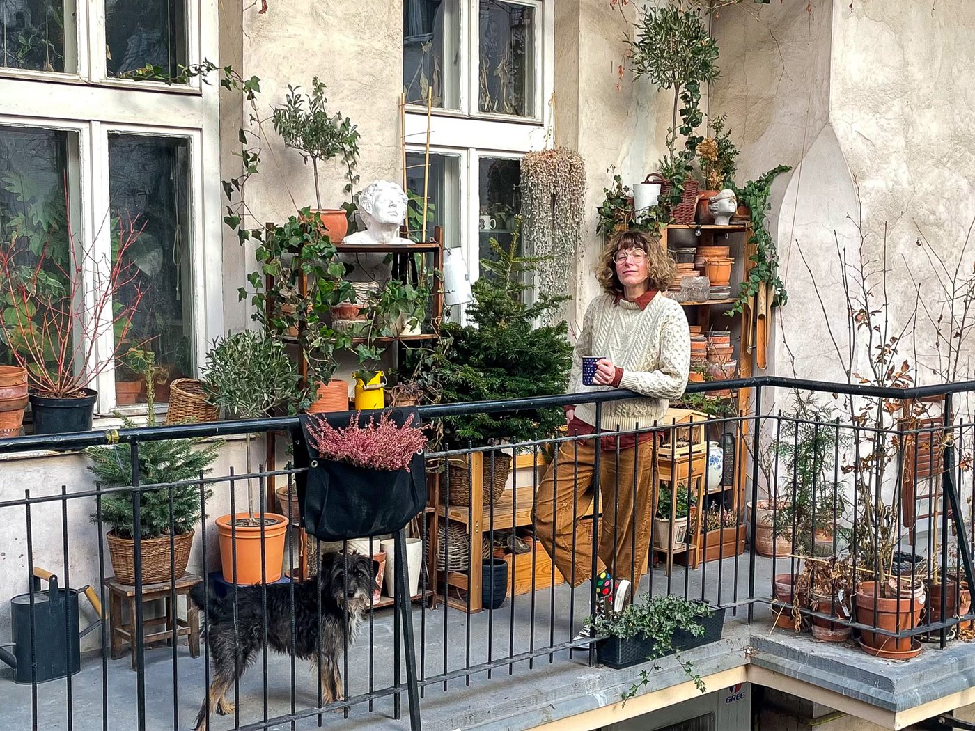 Eine Frau steht mit ihrem Hund auf einem Balkon voller Töpfe, Pflanzen und Gartenutensilien an der Wand.