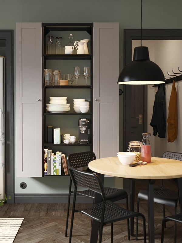 An open shelving unit between two tall cabinets with grey doors, a round wooden table, black chairs and a black pendant lamp.