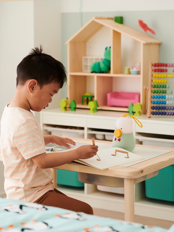 A child sits at a FLISAT children’s table drawing in a book with a green VAPPEBY peanut shape Bluetooth speaker nearby.