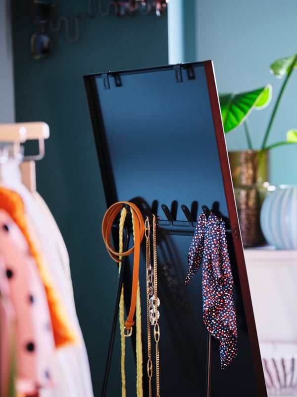 Miroir sur pied KARMSUND noir à côté de vêtements sur cintres. Au dos du miroir, crochets accueillant des ceintures et un foulard.
