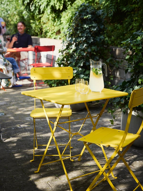 Bright SUNDSÖ tables in red, black, and yellow in a sunny courtyard, with people enjoying their time in the background.
