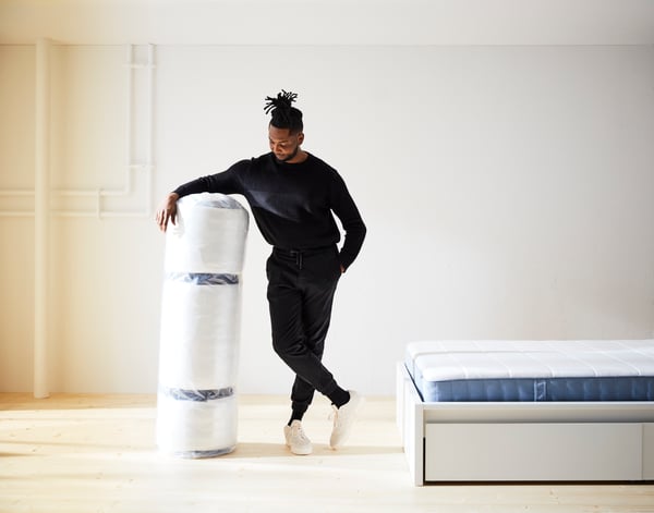 A man holds an extra firm/light blue VESTERÖY pocket spring mattress next to him in a bedroom.