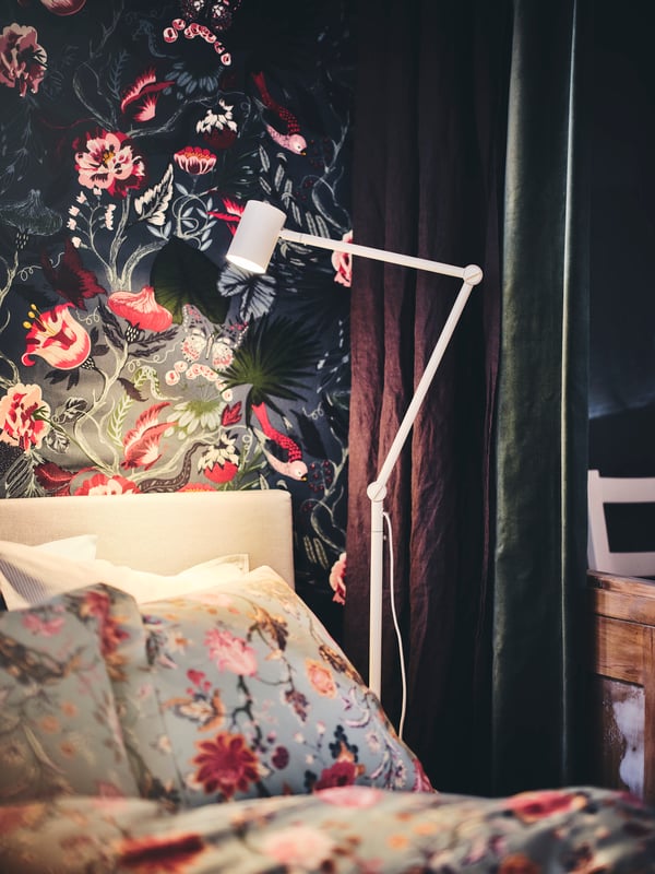 Lampu lantai/bacaan NYMÅNE berwarna putih terletak di sisi katil dengan linen katil NÄSSELKLOCKA yang mempunyai corak berbunga-bunga.