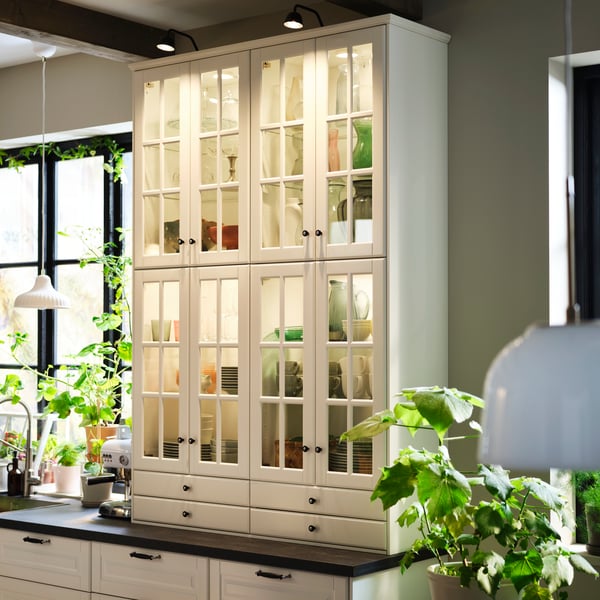 A tall cabinet with off-white BODBYN glass doors showcases kitchenware, with greenery surrounding it in a bright room.
