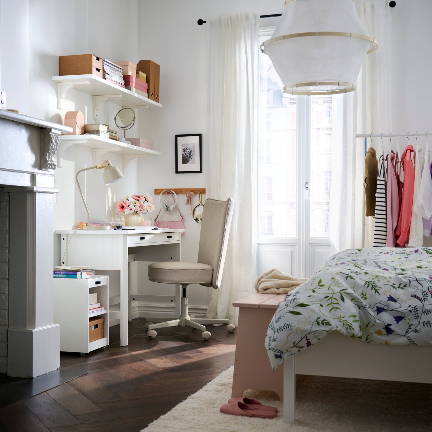 Neben einem Bett mit geblümter Bettwäsche und Kleiderständern steht ein Schlafzimmer mit einem weissen BRYTNING-Schreibtisch aus weissem Material.