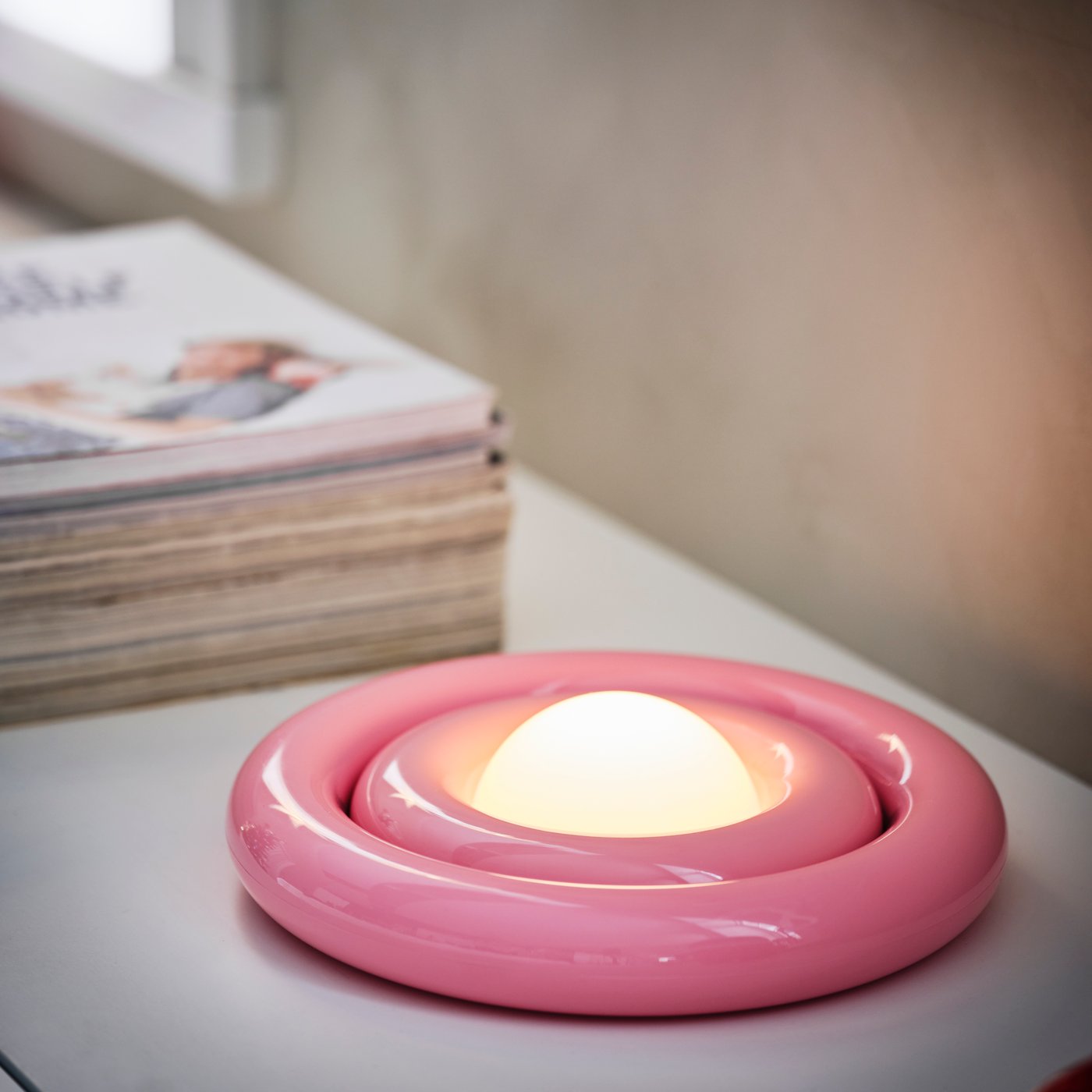 A lit pink STRÅLA LED decorative table lamp is on a white sideboard with a stack of magazines next to it.