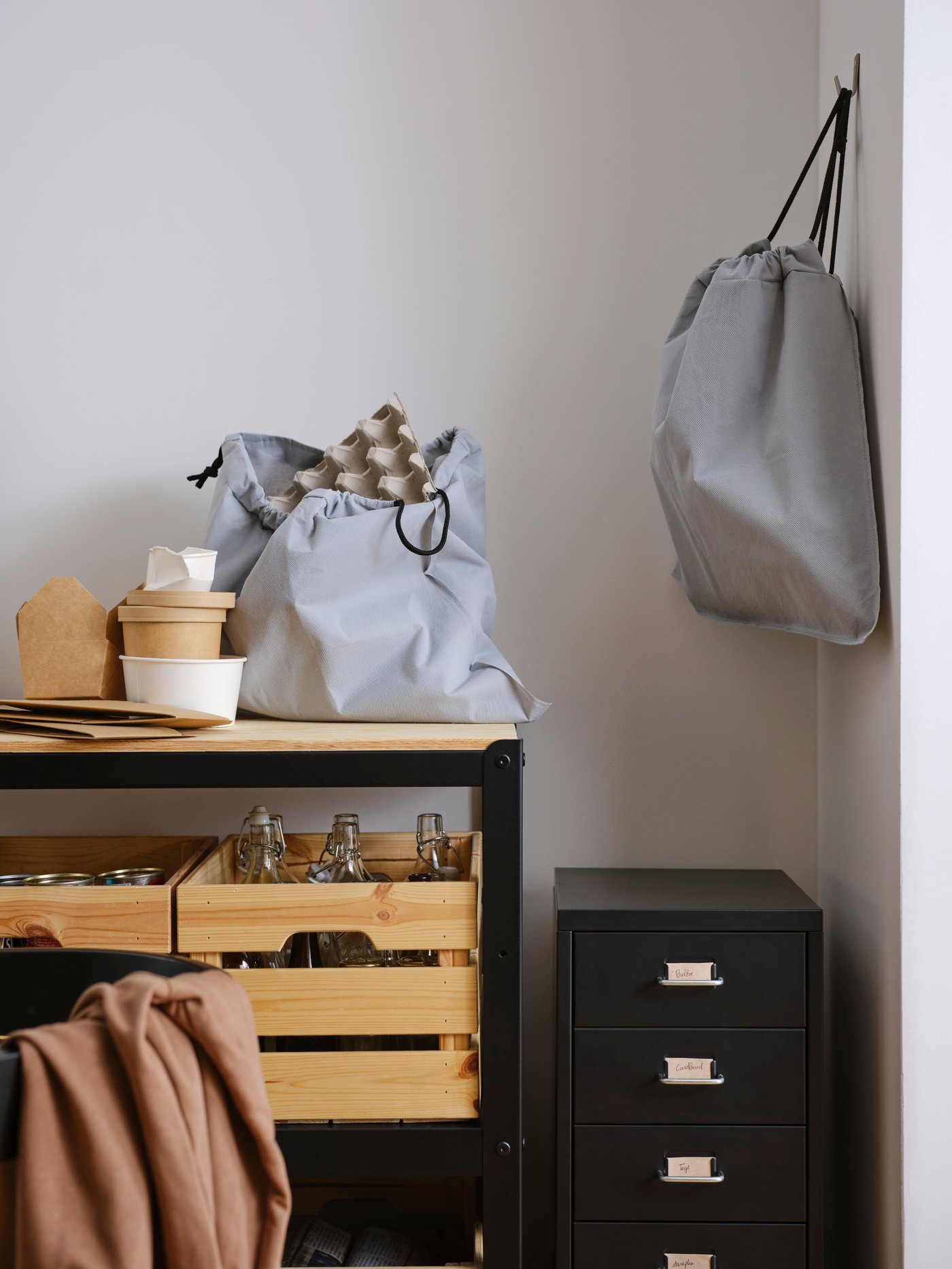 A light grey AJÖSS waste sorting bag on a work bench with pine boxes on a shelf below, plus an identical bag on a wall hook.