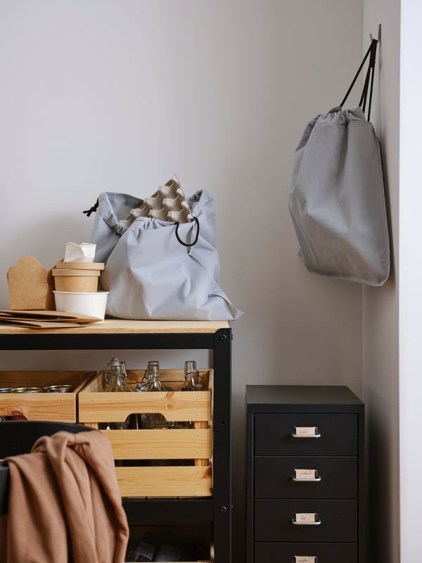 A light grey AJÖSS waste sorting bag on a work bench with pine boxes on a shelf below, plus an identical bag on a wall hook.