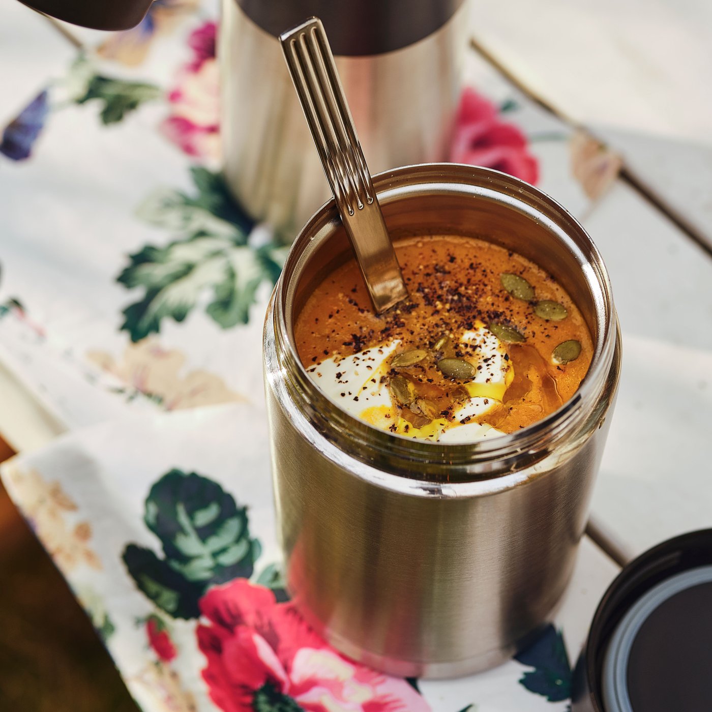 Eine offene EFTERFRÅGAD-Lebensmittel-Isolierflasche mit Suppe und einem Löffel darin sowie ihr Deckel stehen auf einer geblümten Serviette auf einem Tisch.