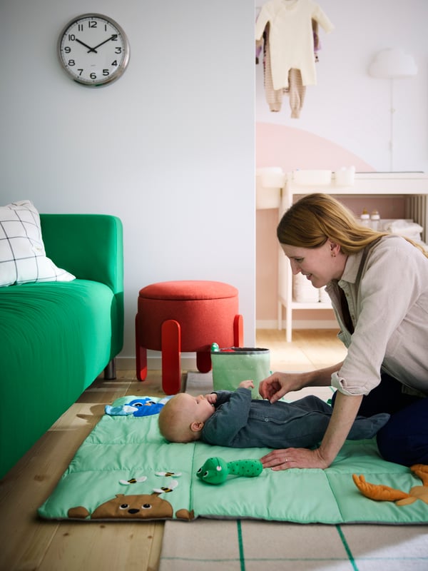 A mom tickling her baby laying on a bright green SANDBI play mat, next to a green sofa and with a red pouffe in the back.