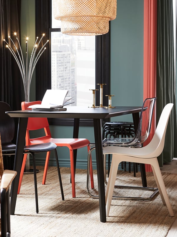 Una mesa de comedor negra rectangular con sillas de diferentes colores como blanco, rojo y negro.