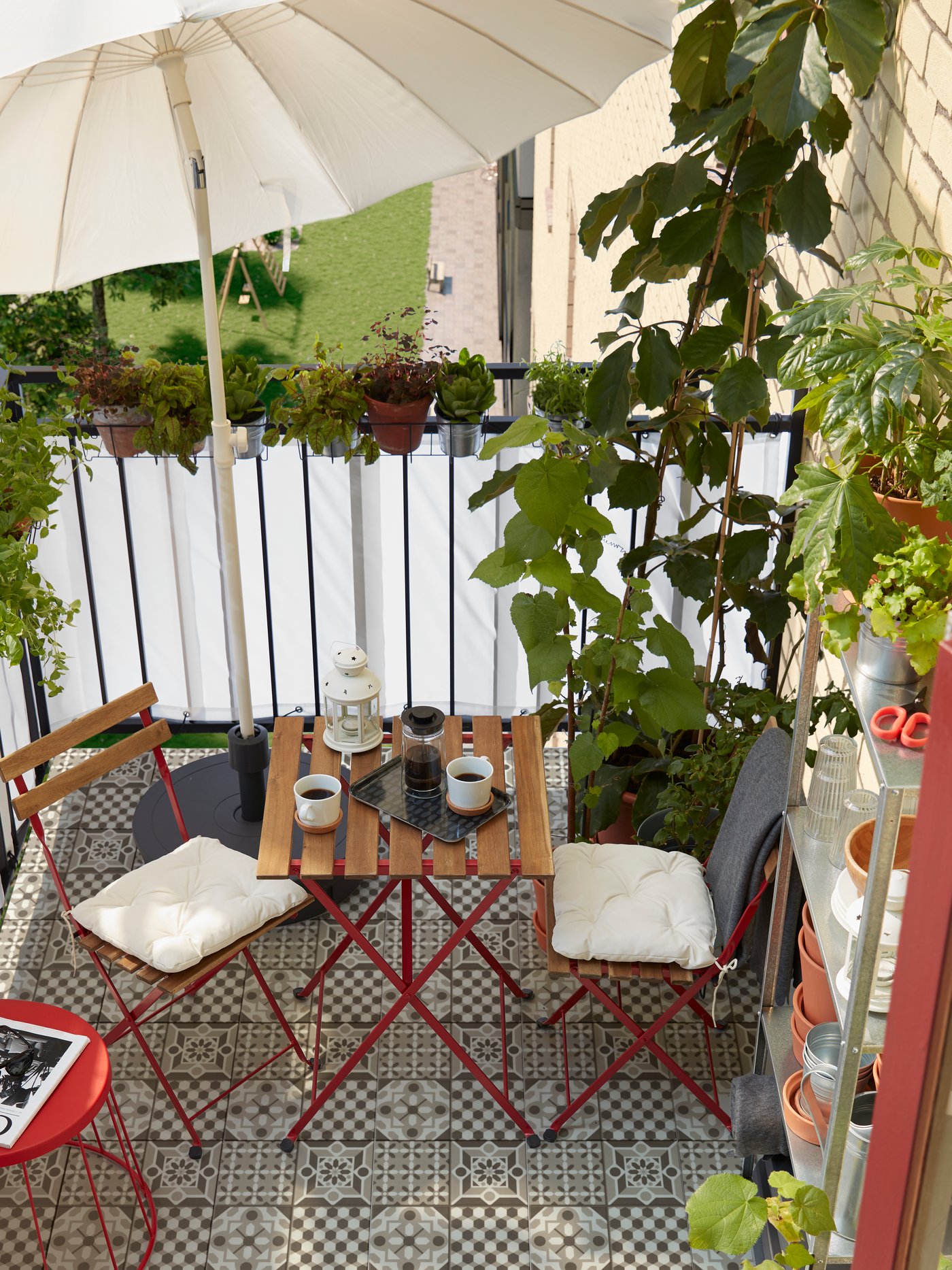 Un pequeño balcón con muchas hierbas y plantas, una estantería con macetas, un parasol, dos sillas y una mesa.