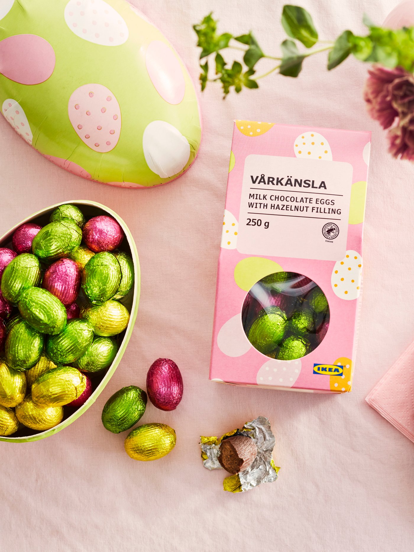 A box of VÅRKÄNSLA milk chocolate eggs on a pink tablecloth, next to an ornate egg filled with colourful milk chocolate eggs.