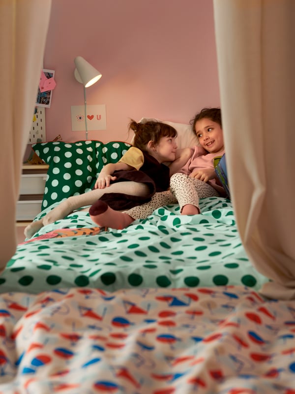 Dos chicas jóvenes relajándose en una cama hecha con un colorido edredón punteado verde colocado en una colorida habitación infantil.