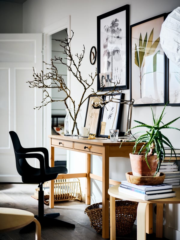 A light brown HEMNES desk against a picture-covered wall, with a vase of branches on top. An ODGER swivel chair is in front.