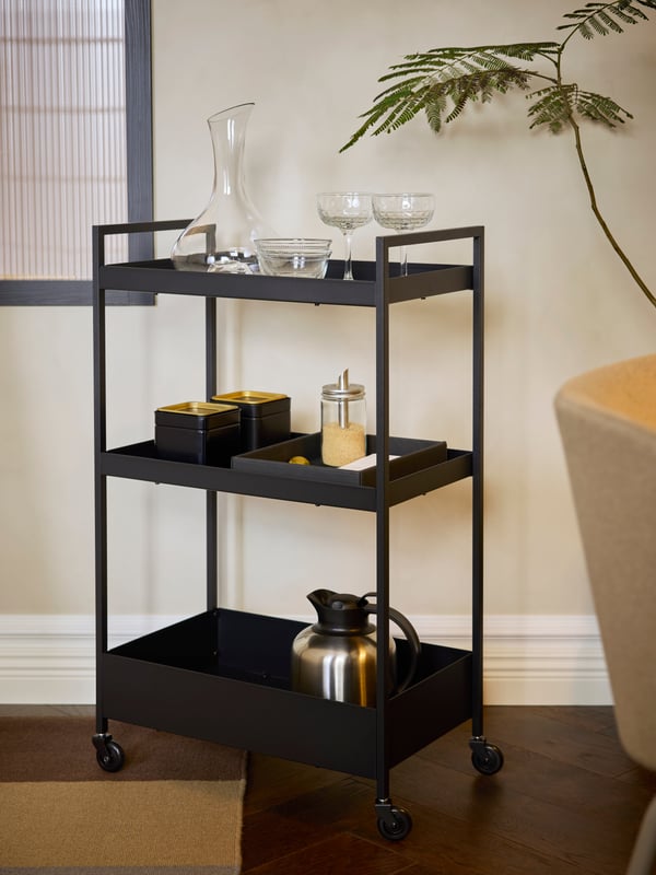 In einem Hotelzimmer steht ein schwarzr NISSAFORS Servierwagen aus Stahl mit drei Böden, auf denen eine Karaffe, Gläser, Teedosen und eine Isolierkanne zu sehen sind.