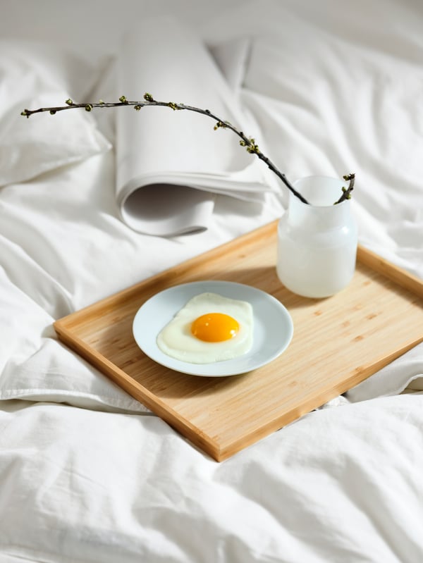 IKEA FÖRENLIG white vase holding flowers, on top of a OSTBIT bamboo tray on top of a bed in the bedroom