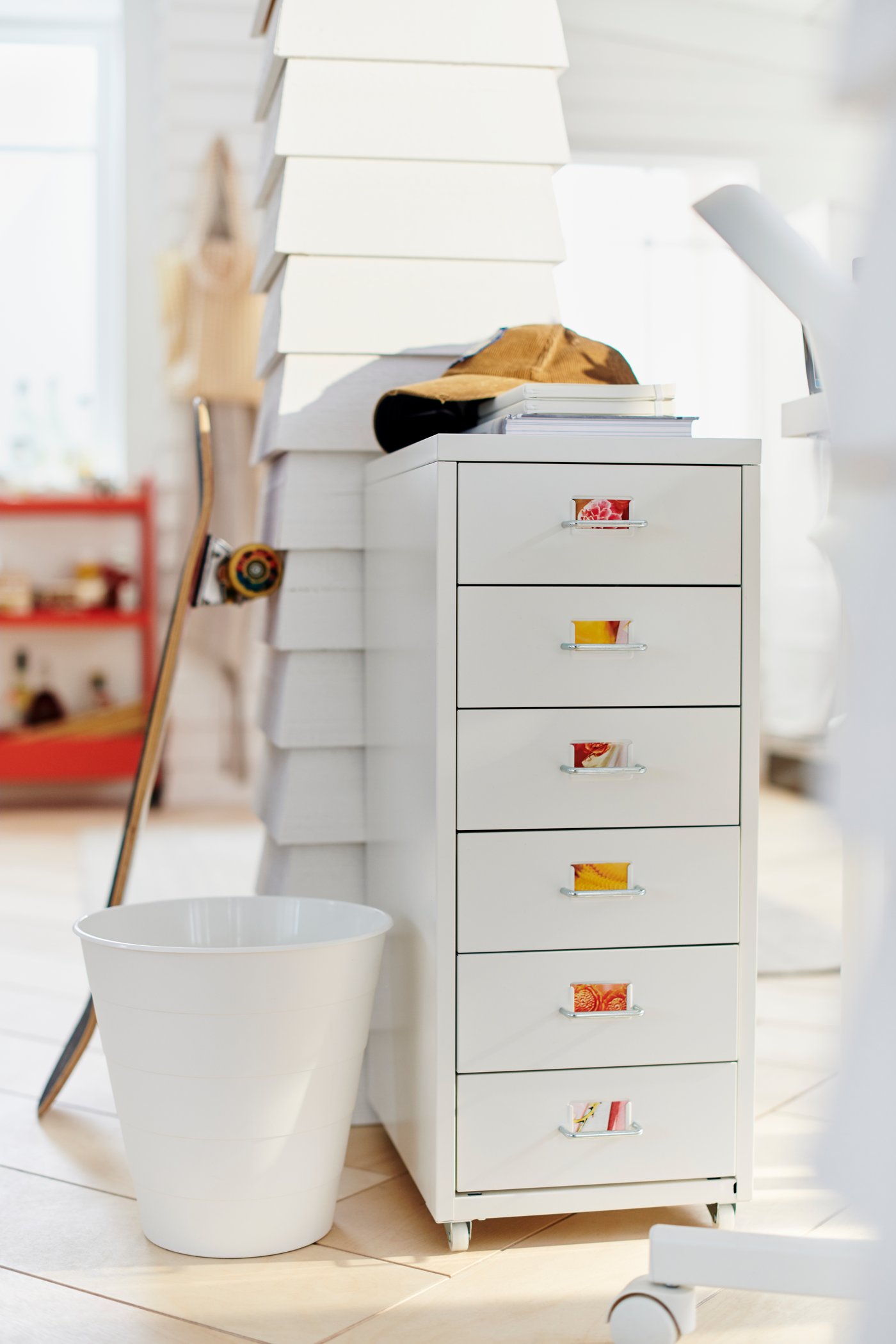 Ein weisses Zimmer, in dem ein HELMER Schubladenelement auf Rollen vor einem getäfelten Holzpfosten steht. An dem Pfosten lehnt ein Skateboard.