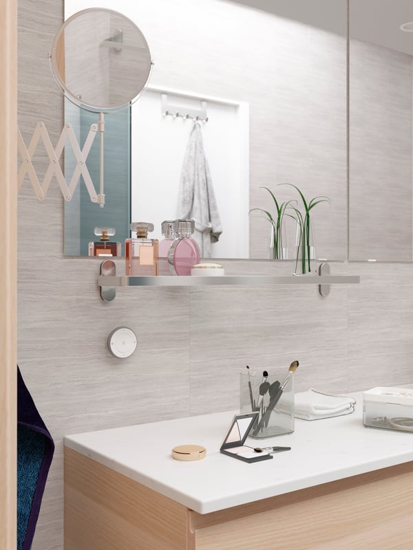 A grey bathroom with a large mirror, a glass BROGRUND shelf underneath with two perfume bottles and a decorative vase.