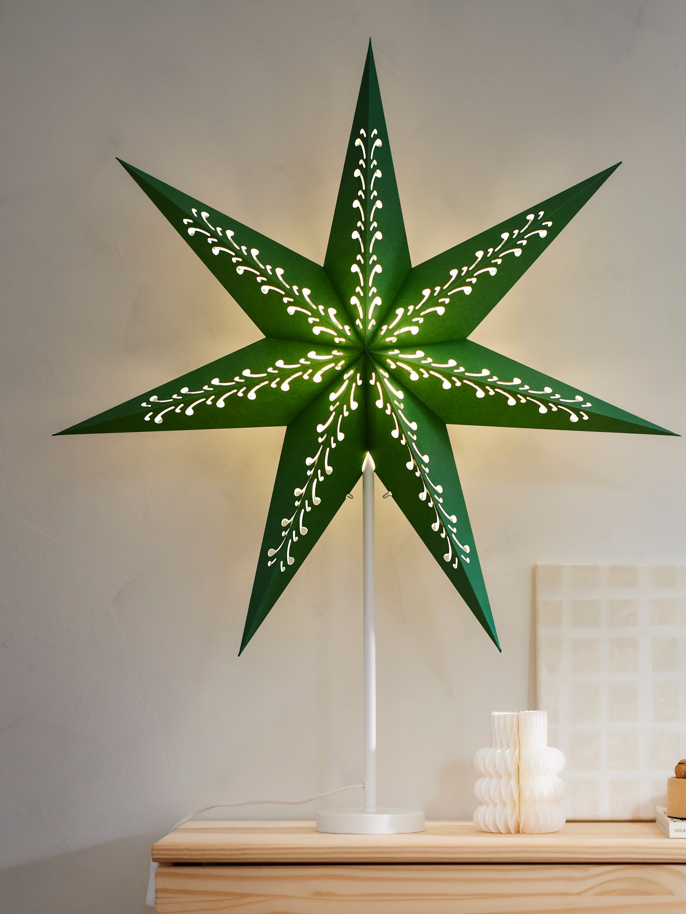 A green glowing STRÅLA star lampshade on white stand on a wooden IVAR shelving unit.