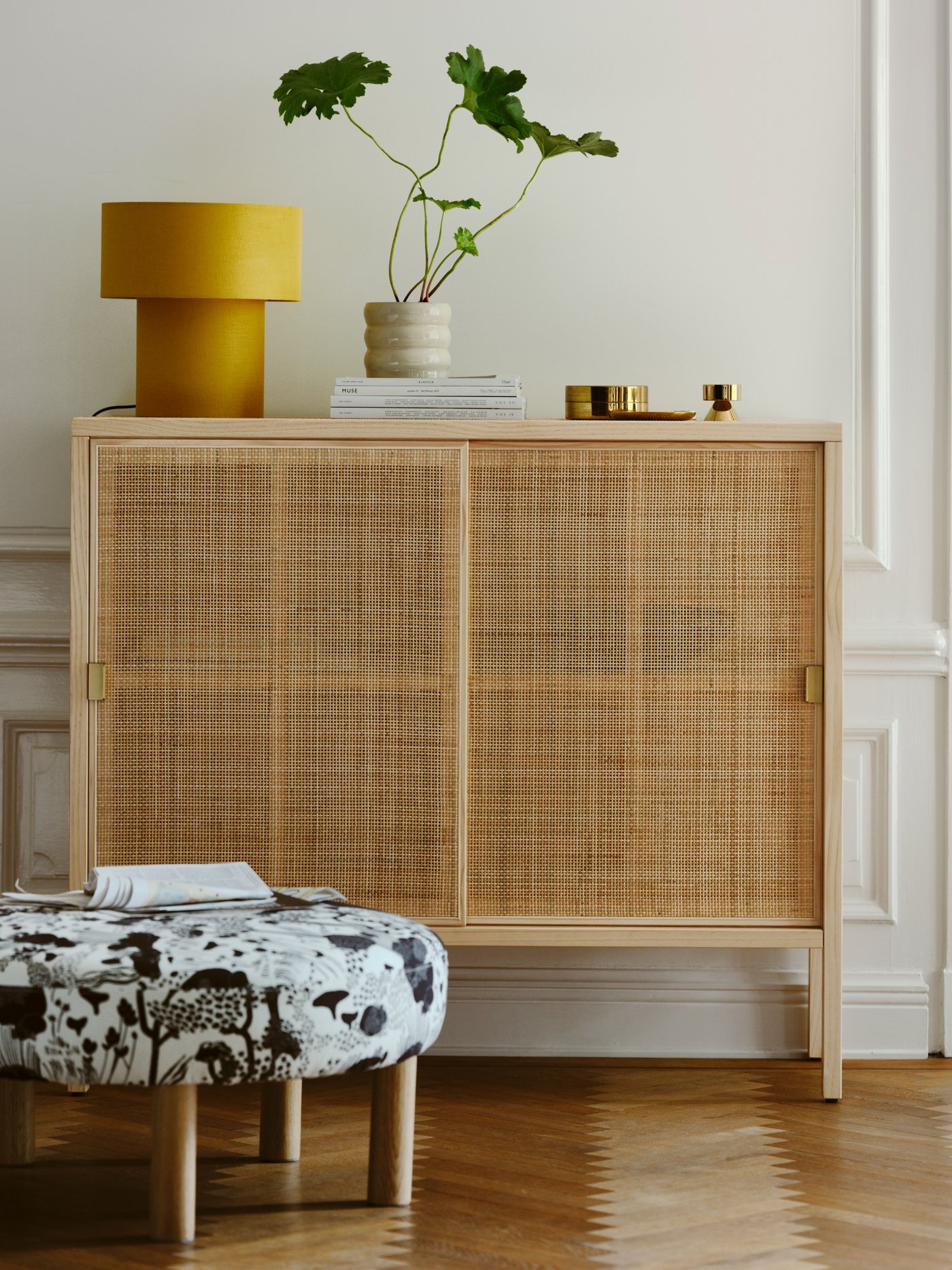 A STOCKHOLM 2025 cabinet with sliding doors in rattan and pine on parquet, with a table lamp on top and a pouffe in front.