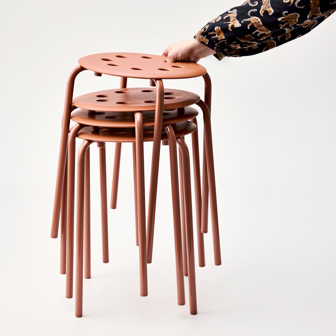 A person is holding a stack of red-brown MARIUS stools, showcasing their lightweight design and versatility.