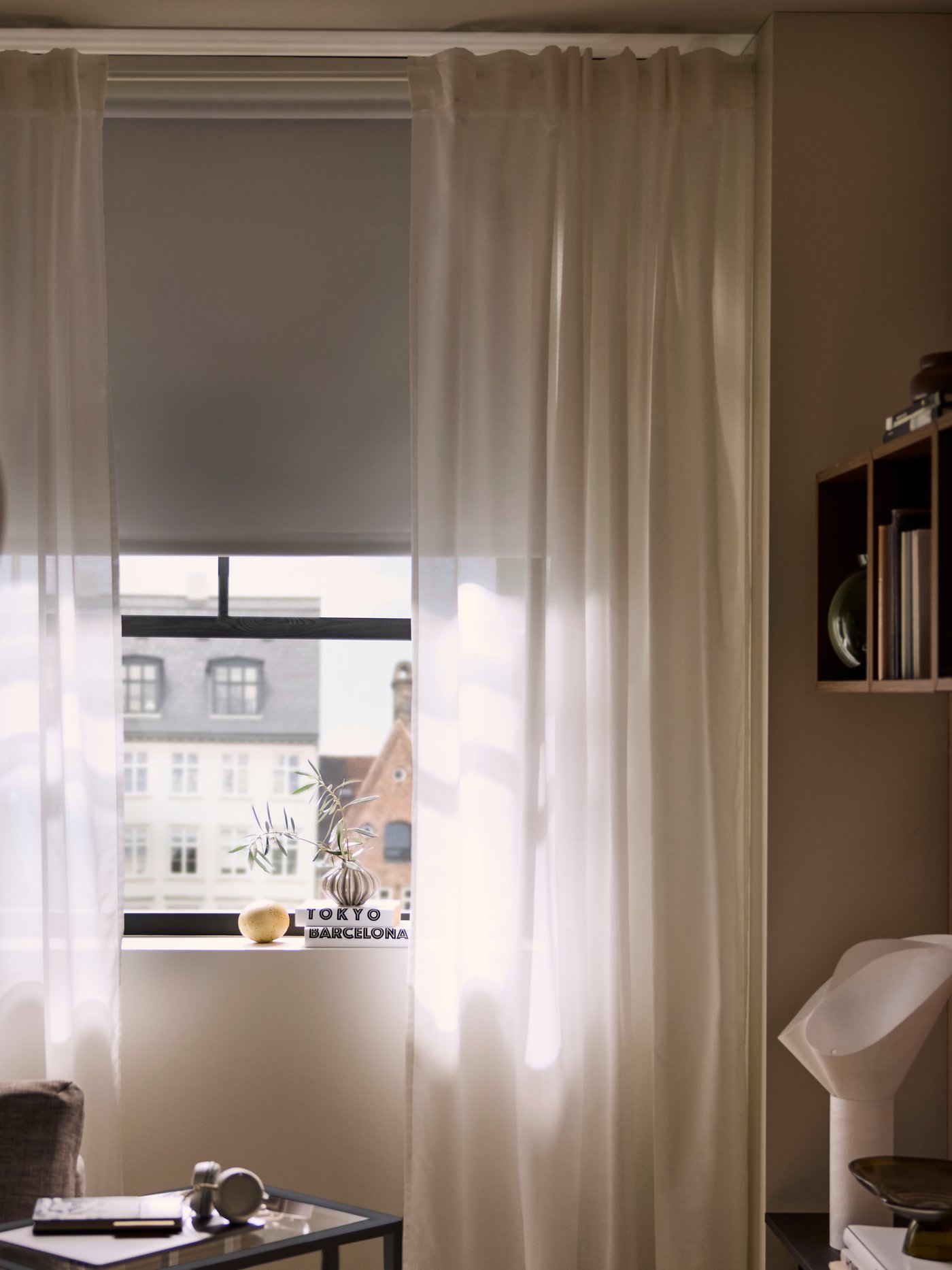 Una ventana iluminada por el sol con cortinas y una persiana FRIDANS de bloqueo de luz parcialmente cerradas para atenuar la luz del interior de la habitación.