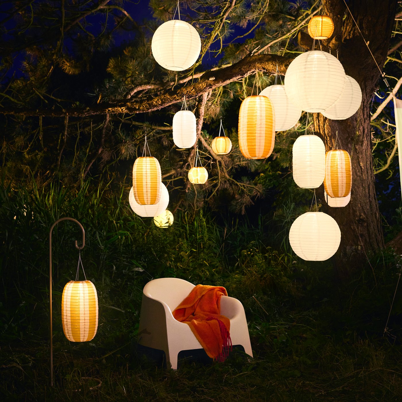 Un jardín al atardecer con una silla blanca y abundantes lámparas colgantes SOLVINDEN de energía solar en tonos blanco y naranja.