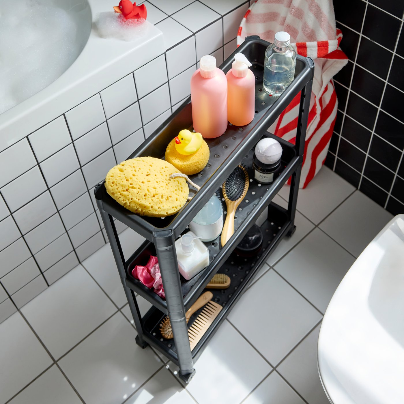 Un carrito VESKEN negro con tres estantes llenos de cepillos para el pelo, botellas de shampú y juguetes para el baño. El piso es blanco.
