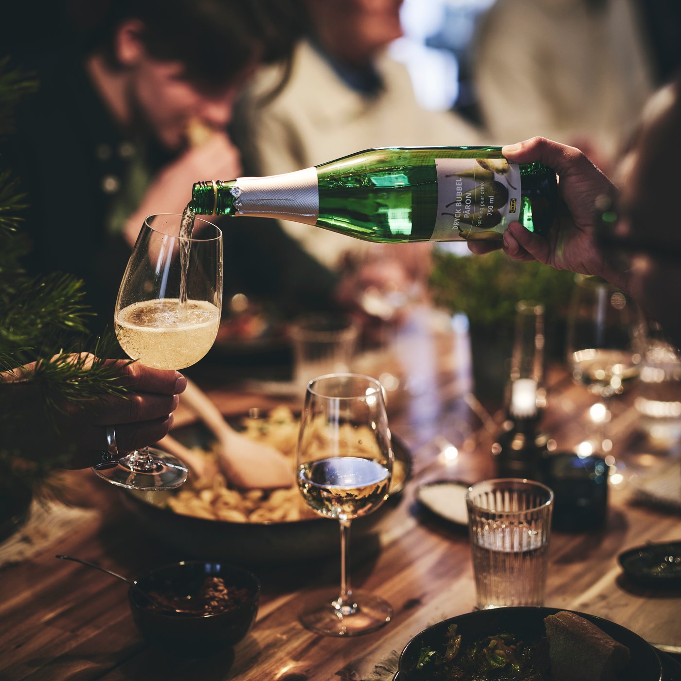 A guest at a dinner party pours a glass of sparkling drink into another guest’s wine glass.
