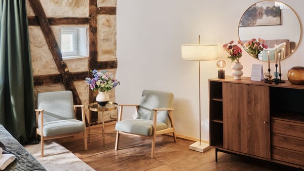 Ett vardagsrum med rustik trävägg och synliga bjälkar, två ljusa fåtöljer med ett litet runt bord mellan, samt en träskänk med lampa, spegel och blommor.