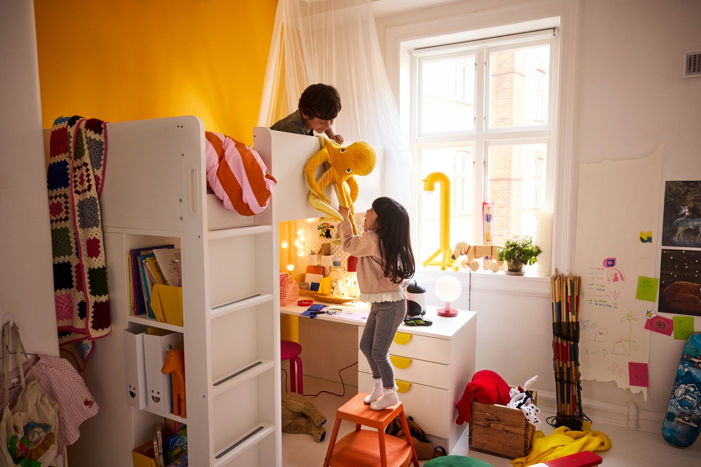 Dans une chambre d’enfants, deux enfants jouant avec une peluche ; l’un est sur le lit en hauteur d’un lit superposé et l’autre est sur un tabouret posé sur le sol.