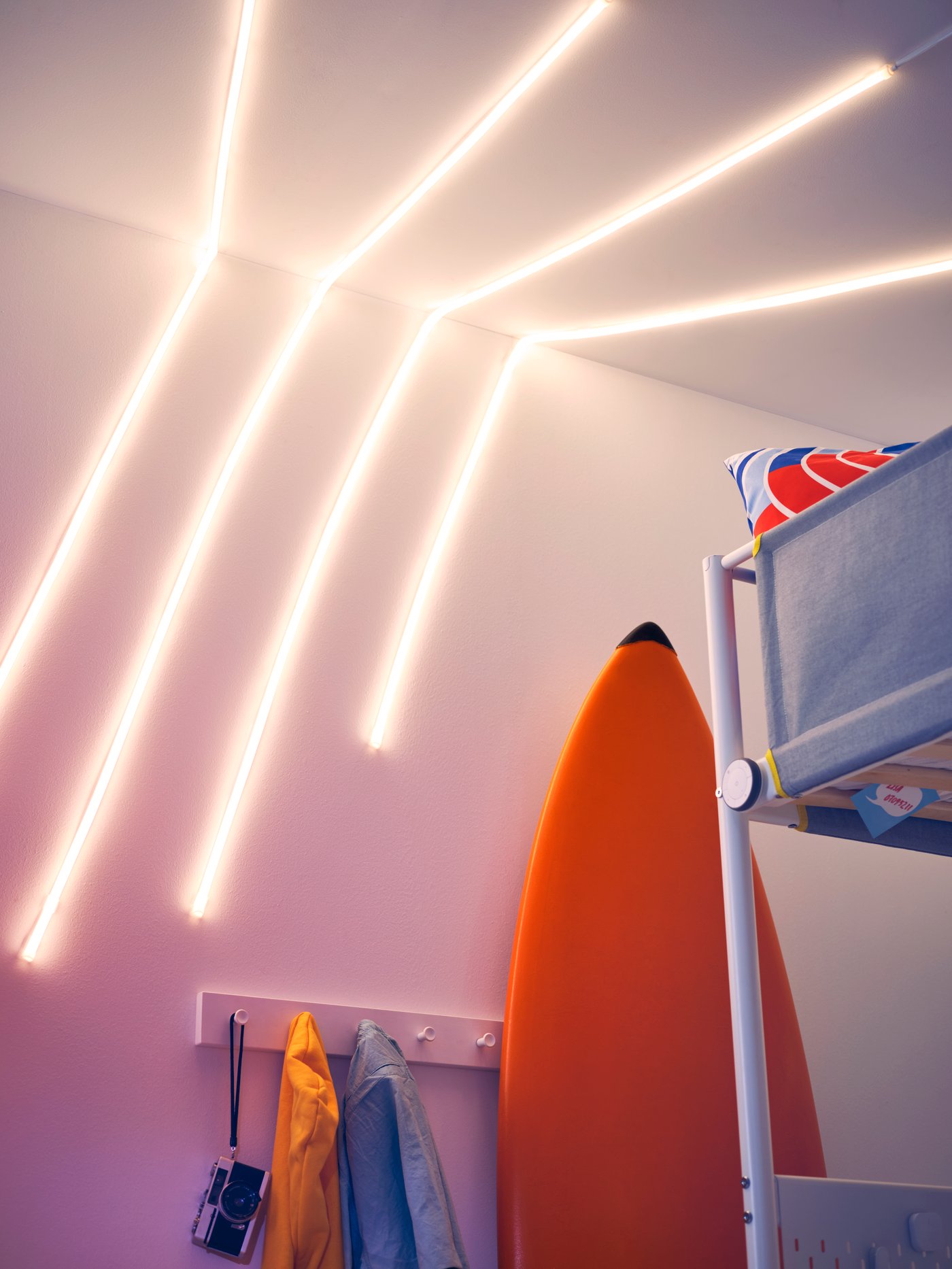 A wall and ceiling with several MYRVARV LED lighting strips, a surfboard, a loft bed, a hook rack with clothes and a camera.