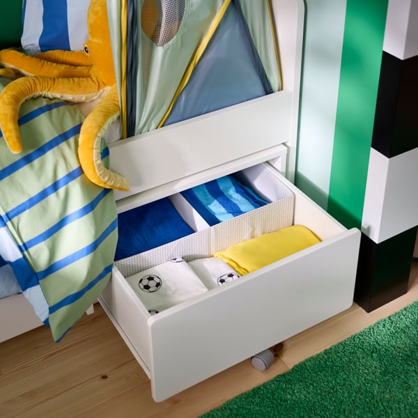 Eine SLÄKT-Aufbewahrungsbox mit Rollen, die unter einem Kinderbett hervorgezogen wurde und mit Bettwäsche gefüllt ist.
