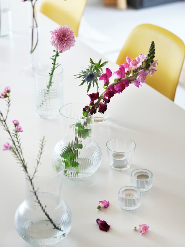 A white tabletop scattered with blossoms, FINSMAK tealight holders and flowers in PÅDRAG clear-glass vases.
