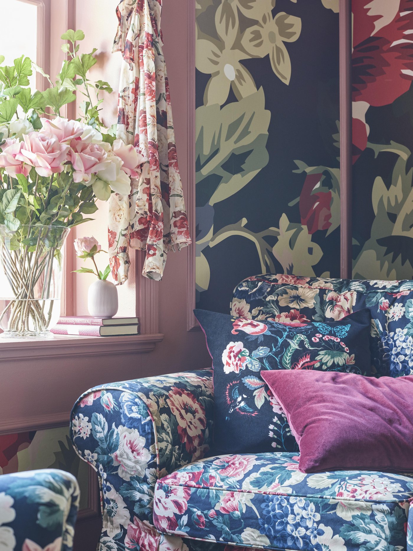 Floral patterned sofa with deep pink cushion in a room decorated with bold floral wallpaper and a pink window frame.