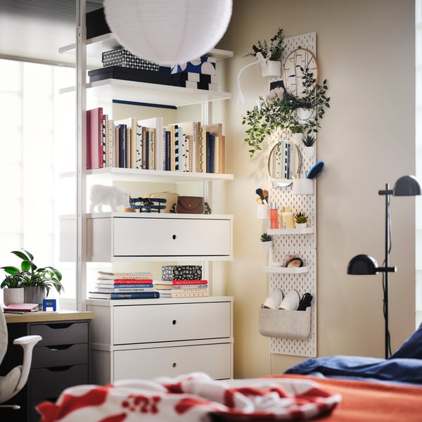 Es ist ein helles Schlafzimmer mit einem weißen ELVARLI Schubladenelement, Böden und Pfosten zu sehen, u. a. mit Büchern und Dekogegenständen. Daneben ist eine Lochplatte platziert.