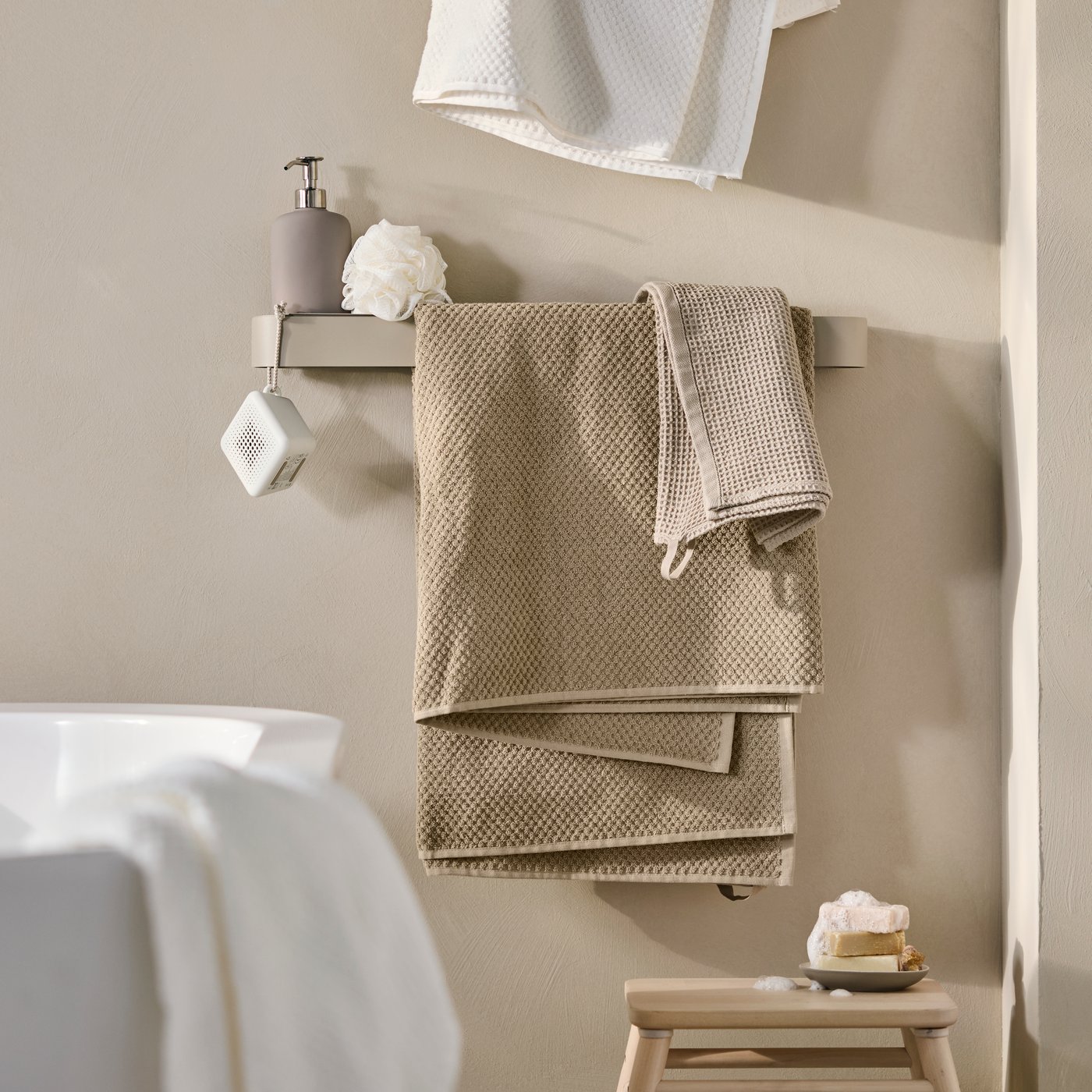 A bathroom with a beige FLORSJÖN towel rail holds folded towels, while a soap dispenser and washcloth are nearby.