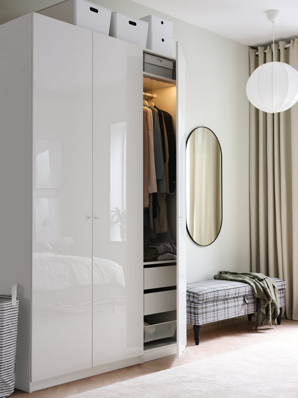 A wardrobe with high-gloss white doors, white storage boxes on top, a bench and a mirror.